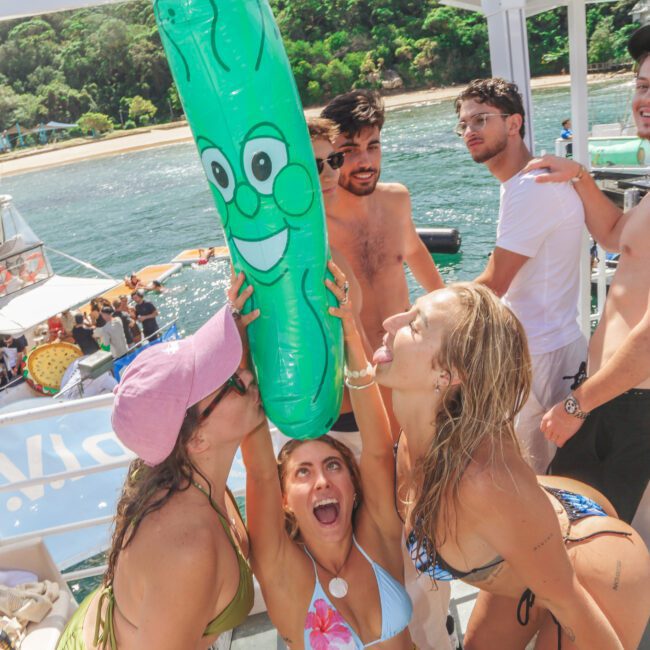 A group of young adults in swimwear enjoy a party on a boat. Three women excitedly pose with a large inflatable pickle, while others smile in the background. The boat is near a shoreline with trees.