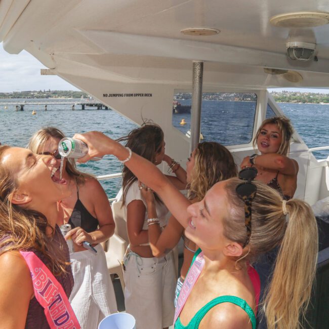 A group of women enjoy a party on a boat. One woman pours a drink into another’s mouth while others watch and laugh. The sea and coastline are visible in the background under sunny skies.