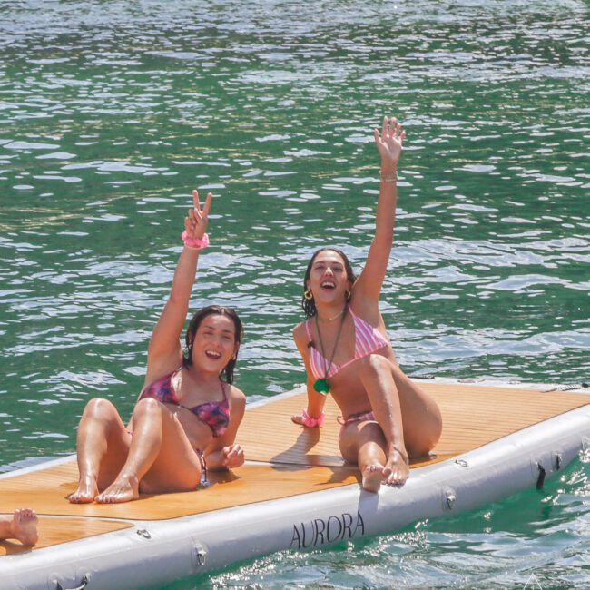 Two women in swimsuits sit on a floating paddleboard in clear, greenish water, both smiling and waving at the camera; one flashes a peace sign, the other raises her hand high.