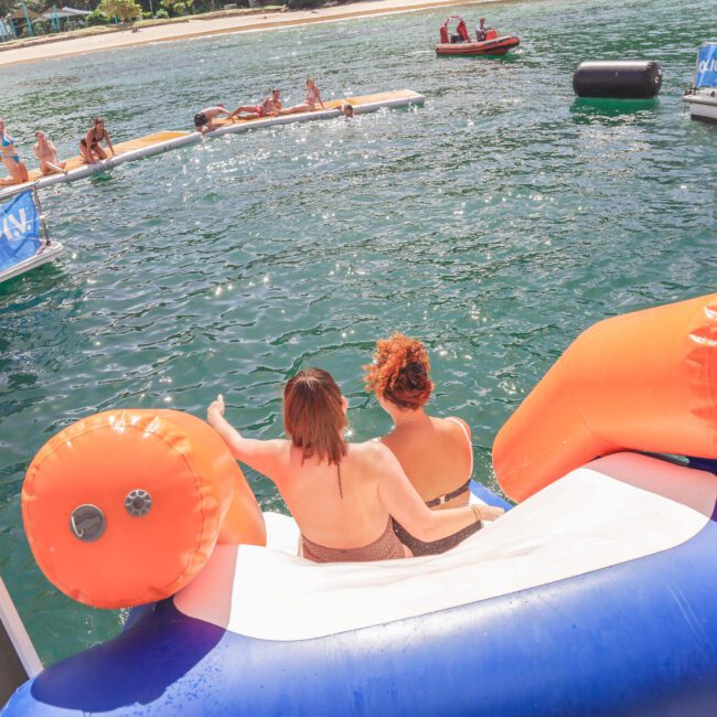 Two women sit at the top of an inflatable slide on a boat, preparing to go into the water. Other people are swimming and relaxing on inflatables nearby under a sunny sky. The scene looks lively and fun.
