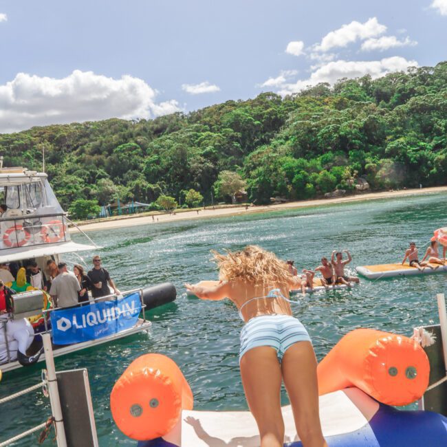 A woman in a swimsuit dives off a yacht into the water, with people and boats in the background near a lush, green shoreline under a partly cloudy sky.