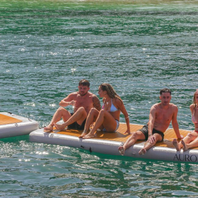 Five young adults in swimsuits sit on floating platforms in the water on a sunny day, smiling and posing for the camera. One person is partly out of frame, appearing to climb onto a platform. The water is green and clear.