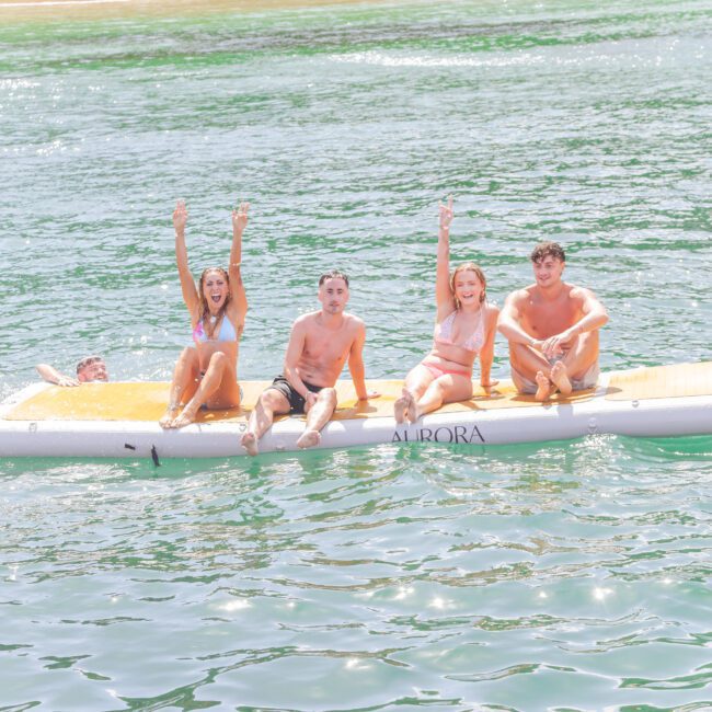 Four people in swimsuits sit and smile on an inflatable float in a river, waving and having fun. A sandy shore and trees are visible in the background under bright sunlight.