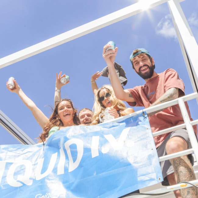 A group of smiling people on a boat raise canned drinks in the air. They stand behind a blue banner and pose under a bright blue sky with sunlight streaming down.