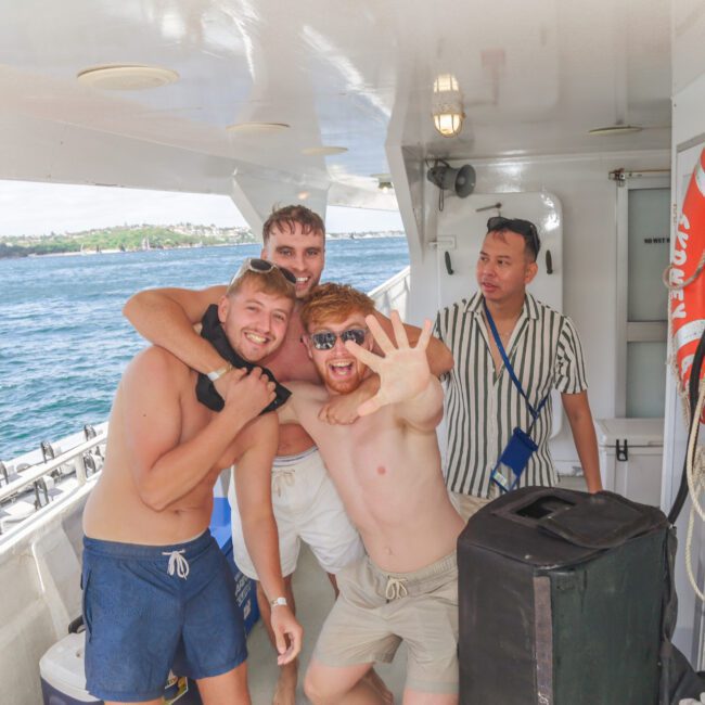 Four men pose playfully on the deck of a boat; three are smiling and laughing together while one stands nearby, looking on. The ocean and coastline are visible in the background. A life preserver hangs on the wall.