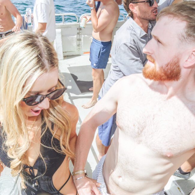 A group of people in swimsuits are on a boat under sunny weather. A woman in sunglasses and a black swimsuit stands next to a shirtless man with a beard. The ocean is visible in the background.