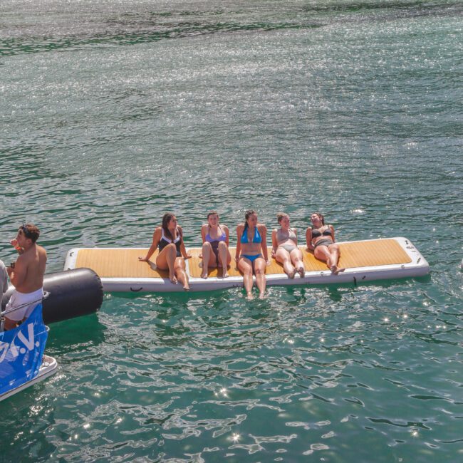 Five people in swimsuits relax on a floating dock in the water near a beach, with trees and sand visible in the background. The sun is shining, and the water sparkles around them. A boat is partially visible on the left.