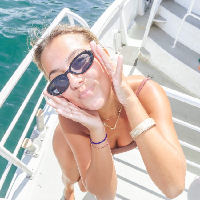 A woman in sunglasses poses playfully on a boat, holding her face with both hands and smiling. The sunny day highlights the blue water and white railings in the background.
