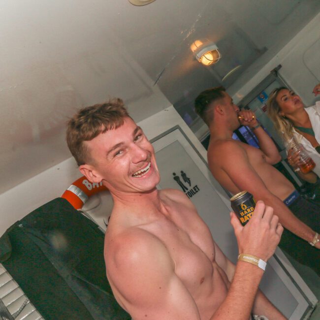 A shirtless man smiling and holding a drink can at a lively indoor party with other people drinking and socializing in the background. The atmosphere is casual and festive.