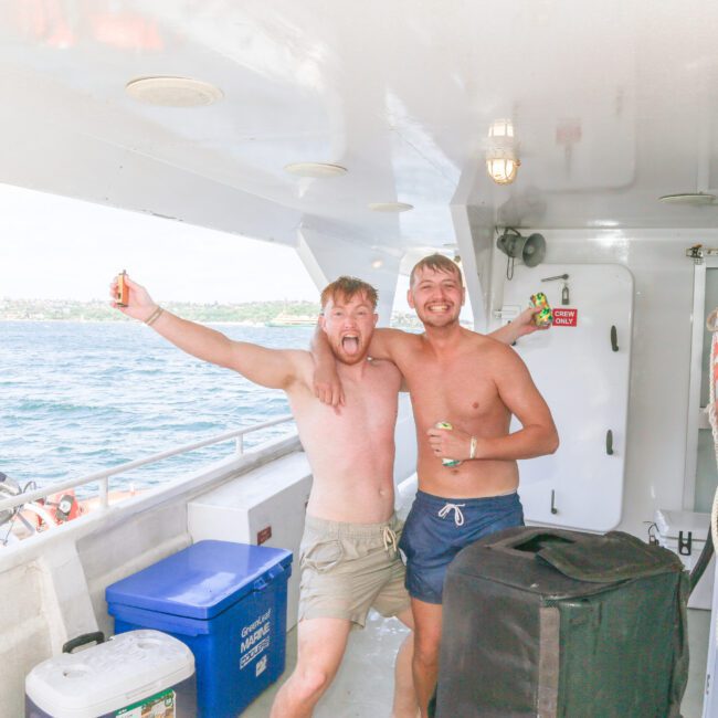 Two shirtless men smiling and hugging on the deck of a boat, each holding a drink. The ocean and coastline are visible in the background, along with life rings and coolers on the boat.