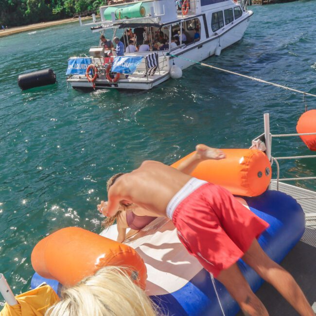 A man in red swim trunks leans over an orange and white inflatable raft on a dock, with a boat full of people on the water in the background. Another man with a towel and drink stands nearby.