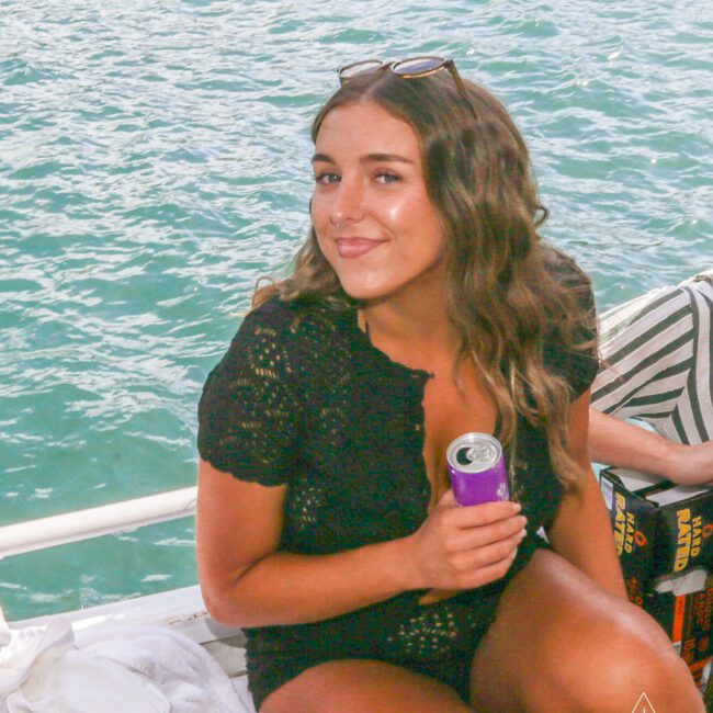 A woman with long wavy hair, wearing a black outfit and sunglasses on her head, sits on a boat holding a purple can, smiling at the camera with water in the background.