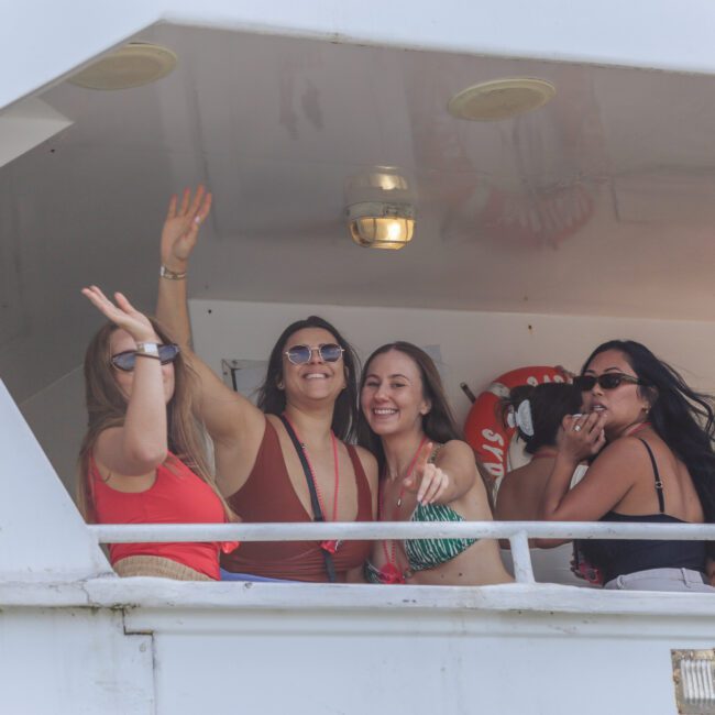 Four women stand on a boat deck, smiling and waving. They are dressed in summer attire and appear to be enjoying themselves. Bright light suggests daytime, and a life preserver is visible in the background.