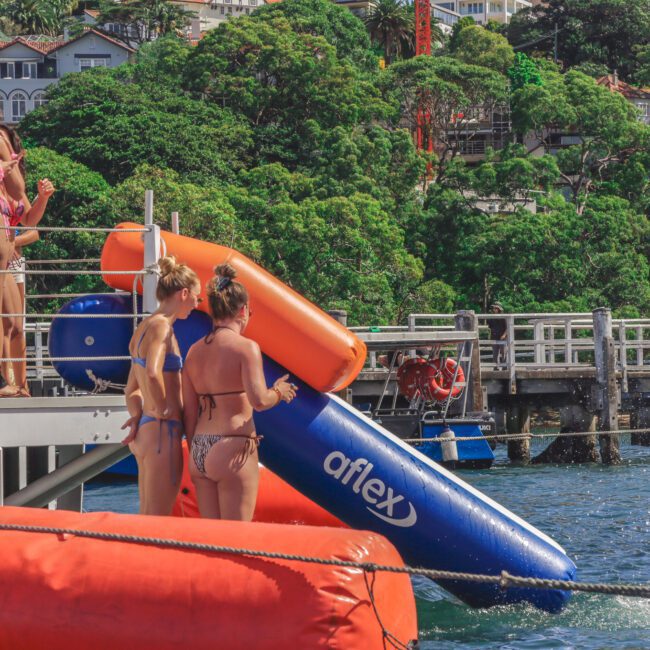 Several people stand on a dock next to an inflatable blue and orange water slide labeled "aflex," with lush greenery and houses visible in the background. Some are in swimsuits, ready to use the slide.