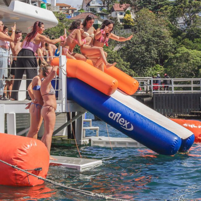A group of people in swimsuits laugh and slide down two inflatable slides from a boat into the water, while others watch and cheer. Houses and trees are visible in the background.