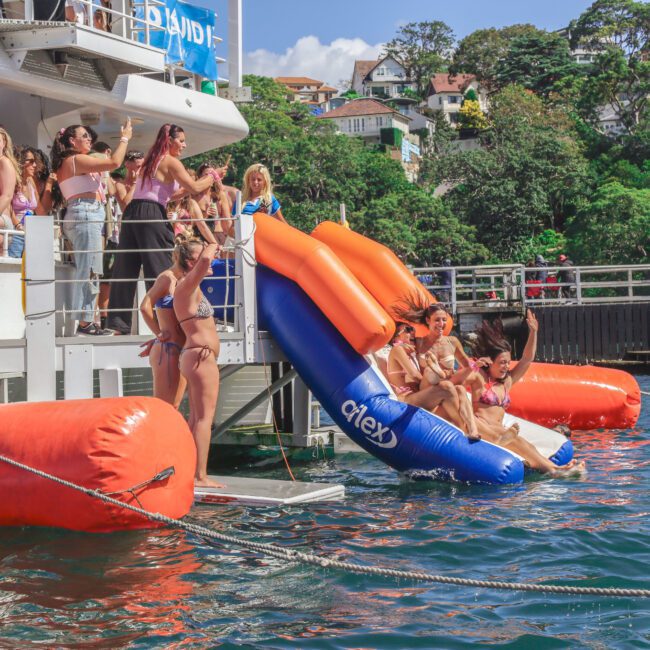 A group of women in swimsuits slide down an inflatable slide from a boat into the water, while others watch and enjoy the sunny day. Houses and lush trees are visible on the hillside in the background.