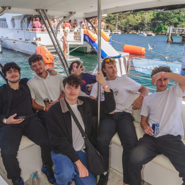 Five young men are sitting and relaxing on a boat, some holding drinks, with water, other people, and boats visible in the background. The atmosphere appears casual and social.