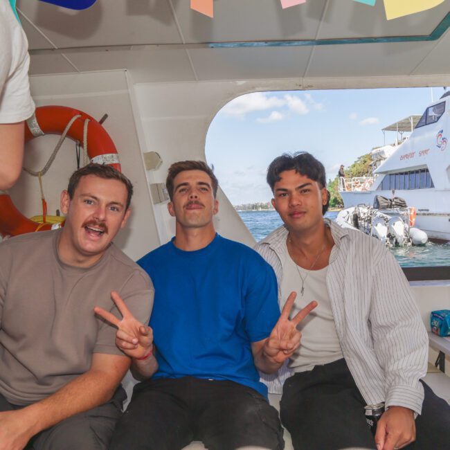 Three young men sit and smile on a boat, two of them holding up peace signs. There are snacks and drinks on a table nearby, and another boat, water, and trees are visible through the window behind them.