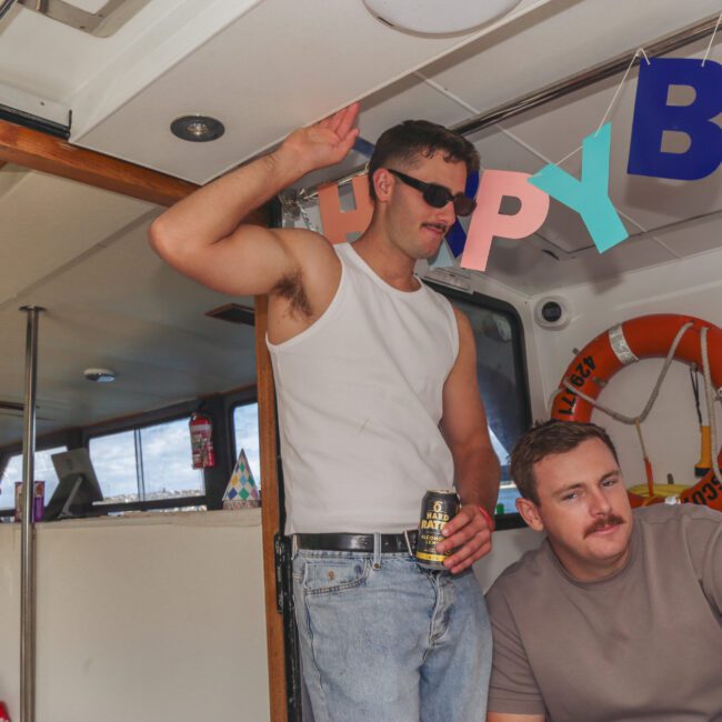 A man in sunglasses and a white tank top stands holding a drink and raising his hand, surrounded by three other men on a boat decorated with colorful "HAPPY BIRTHDAY" letters. All appear to be enjoying a celebration.