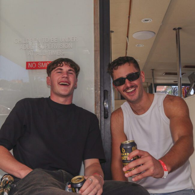 Two young men sit and laugh, each holding a can of drink. They are on a boat, and a fire extinguisher and a safety sign are visible behind them. It's sunny, and both men look relaxed and happy.