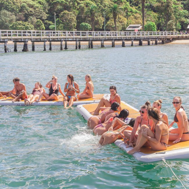 A group of people in swimsuits relax and socialize on floating mats in the water near a wooden pier, with trees and houses in the background under a sunny sky.