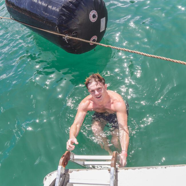 A man in swim trunks climbs a ladder from turquoise water onto a boat, with a large black buoy floating nearby. Sunlight reflects off the water.