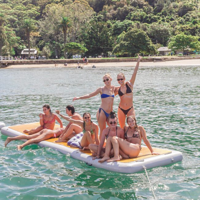 A group of six people in swimwear relax and pose on a floating platform in calm, green water near a lush, forested shoreline, enjoying a sunny day.
