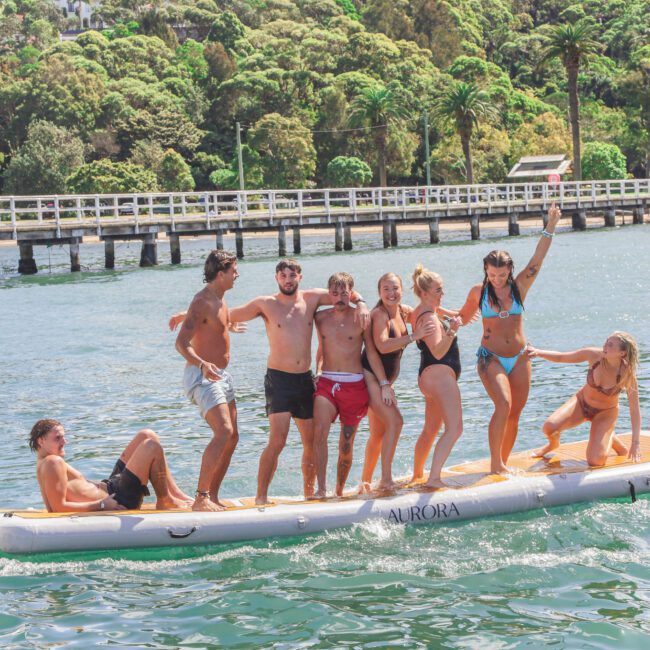 A group of eight young adults in swimsuits balance and have fun on a large inflatable paddleboard on the water, with trees, a white bridge, and a few houses visible in the sunny background.