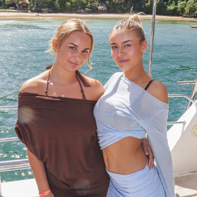 Two women standing on a boat, posing and smiling, with calm blue water and a sandy, tree-lined shoreline in the background on a sunny day.