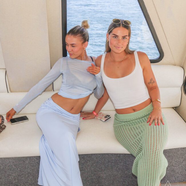 Two women sit on a white cushioned bench inside a boat. One wears a sheer blue top and matching skirt, posing with her arm up, while the other wears a white tank top and green crocheted skirt. Water and coastline are visible through the window.