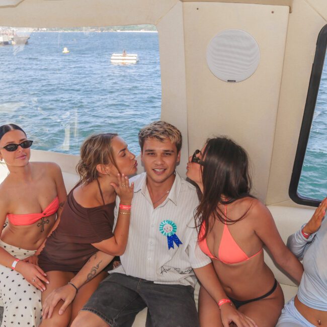 A young man wearing a birthday ribbon sits on a boat, smiling, as two women in swimwear kiss his cheeks and two others sit beside him. The ocean and other boats are visible through the windows.