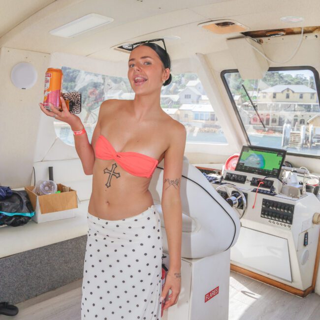 A woman in a pink bikini top and white polka-dot skirt holds a drink and poses inside a boat. She has tattoos, a necklace, and wears sunglasses on her head, with water and buildings visible through the windows.