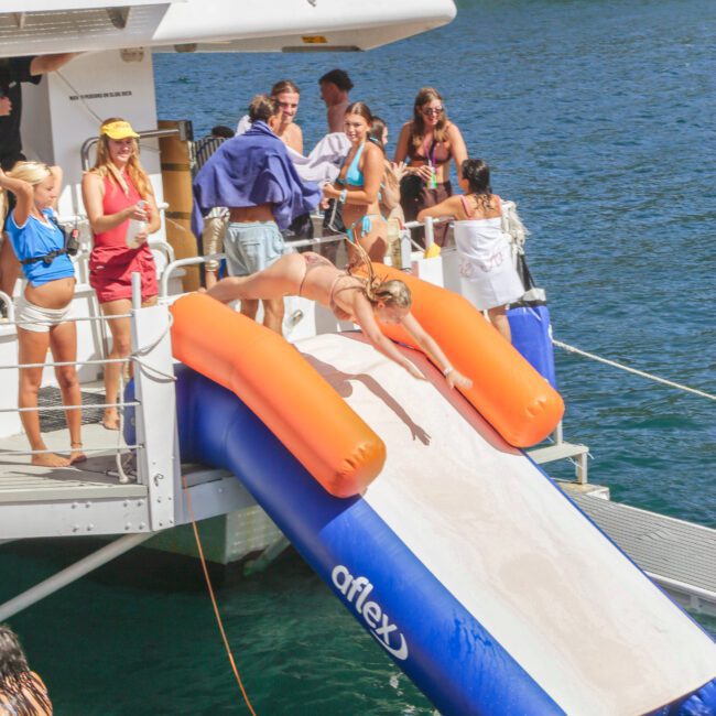 A woman slides headfirst down an inflatable slide from a boat into the water, surrounded by people in swimsuits enjoying a sunny day and watching from the boat.