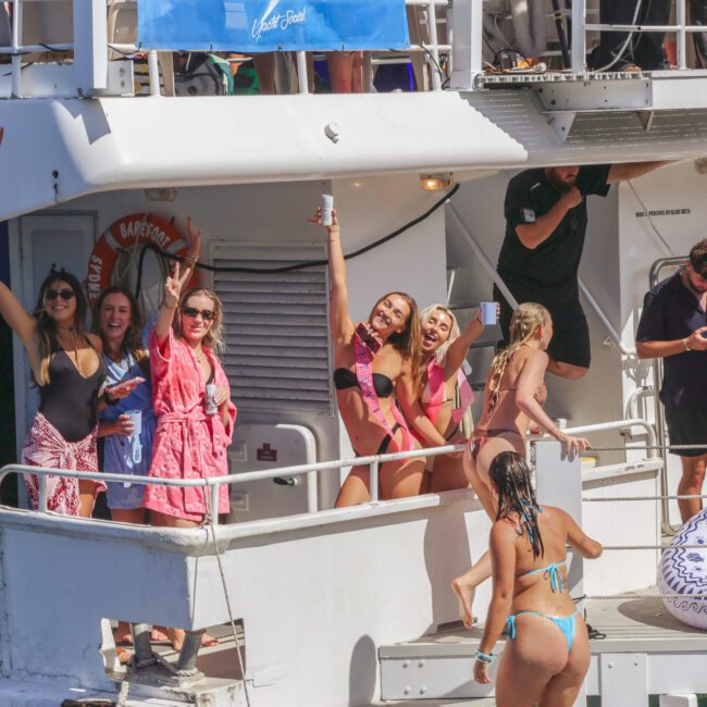 A group of people in swimsuits and robes cheer and pose on the deck of a boat, with some holding drinks, enjoying a sunny day by the water. An inflatable pool float is on the lower deck.