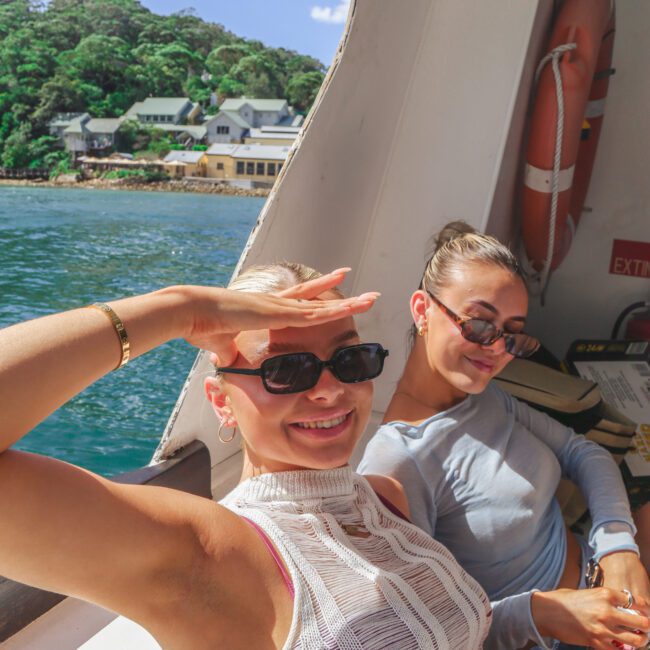 Two women sit and relax on a boat, smiling at the camera. One is shading her eyes from the sun with her hand, while the other sits beside her, both wearing sunglasses. Water and buildings on shore are visible in the background.