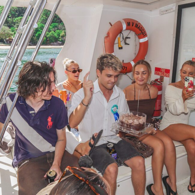 A group of young adults celebrate on a boat. One person holds a birthday cake, surrounded by friends, some clapping and taking photos. Trees and water are visible through the boat's open side. It's a sunny, cheerful scene.