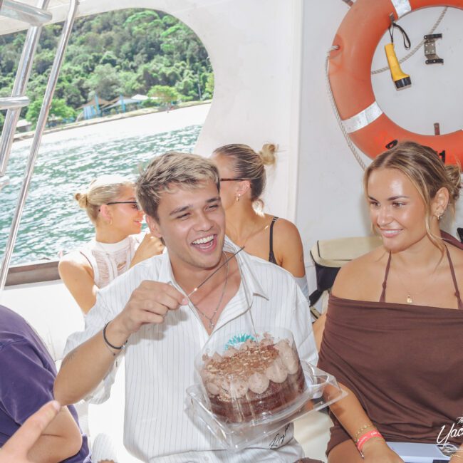 A group of young people are smiling and celebrating on a boat. One person holds a chocolate cake, and the ocean and a lush, green shoreline are visible in the background. A life preserver hangs on the wall behind them.