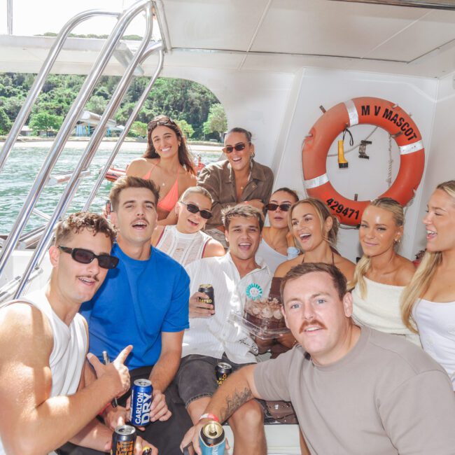 A group of twelve young adults smiling and posing together on a boat, with drinks in hand. They're sitting and standing under a canopy, with water and greenery visible in the background. The atmosphere is lively and joyful.