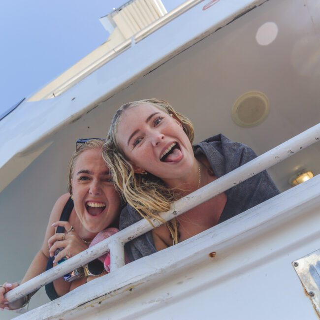 Two smiling people leaning over the white railing of a boat, both sticking out their tongues and looking at the camera under a clear blue sky.