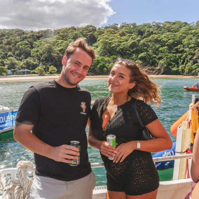 A man and a woman stand on a boat holding drinks, smiling at the camera. Lush green trees and water are visible in the background. The sky is partly cloudy. The words "Yacht Social" are visible in the corner.