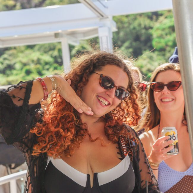 Two women wearing sunglasses smile and pose for the camera at an outdoor event, one holding a drink can and the other making a playful hand gesture near her face. Bright sunlight and greenery are visible in the background.