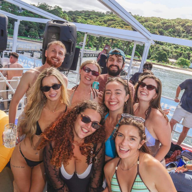 A group of smiling young adults in swimsuits pose together on a boat, with trees and water visible in the background on a sunny day.