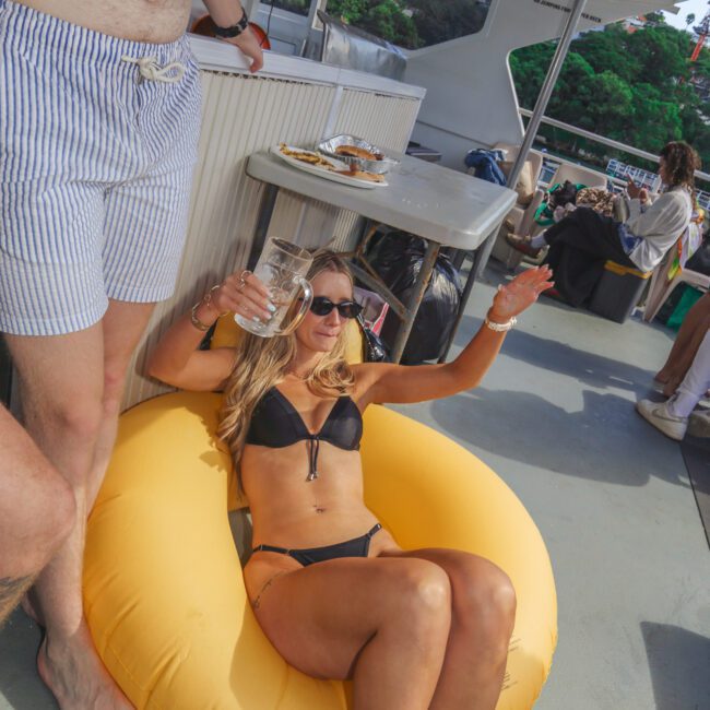 A woman in a black bikini relaxes on a yellow pool float, holding a drink and raising her arm on a boat. Another person stands nearby in striped shorts. Other people and drinks are visible in the background.