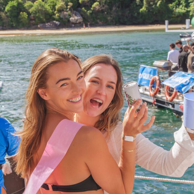 Two young women smile and pose together on a boat. One wears a black bikini and a pink sash, the other wears a white dress and holds a can. They are surrounded by water and lush greenery in the background.