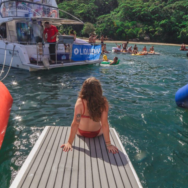 A woman in a red swimsuit sits at the edge of a dock extending into the water, facing a boat with people onboard and others swimming nearby. Lush green trees and a blue sky are in the background.