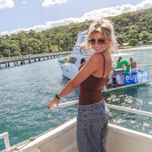 A woman in sunglasses and casual clothes stands on a boat, smiling at the camera. Behind her, a dock and another boat with people on it are visible, surrounded by blue water and green trees under a sunny sky.