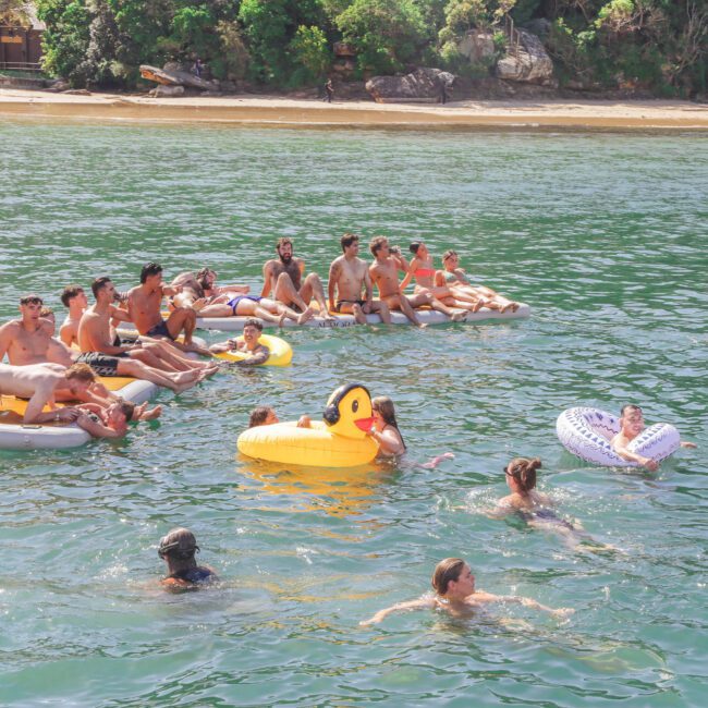 A group of people relax on inflatable rafts and floaties, including a large yellow duck, in a sunny, green-blue body of water near a sandy beach with trees in the background.