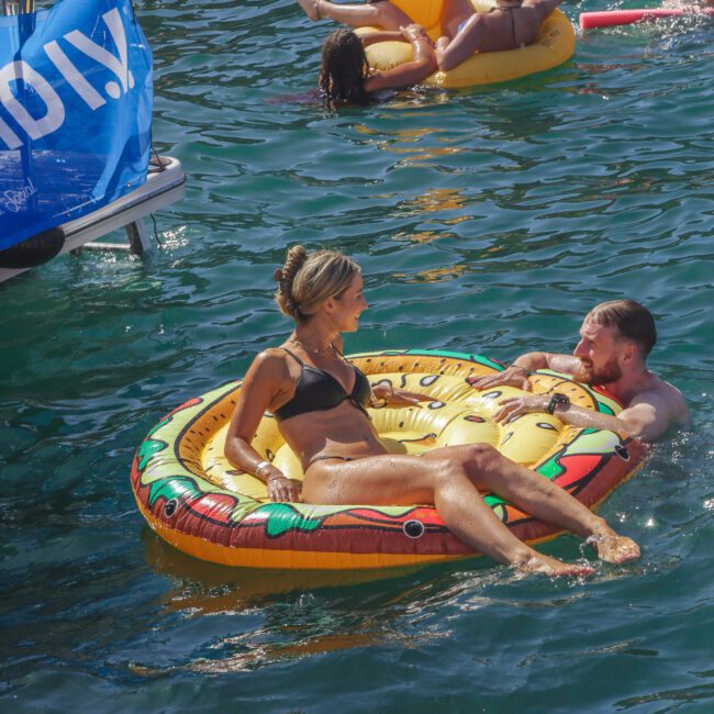 A woman in a black bikini lounges on a pizza-shaped float while a man holds on beside her in the water. Other people are relaxing on inflatables nearby in a sunny, lively outdoor setting.