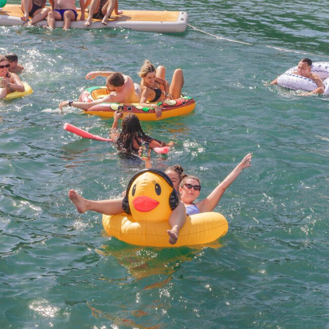 People relax and have fun on inflatable pool floats in clear blue water. Two women smile on a yellow duck float in the foreground, while others float and sit together in the background. The mood is playful and joyful.
