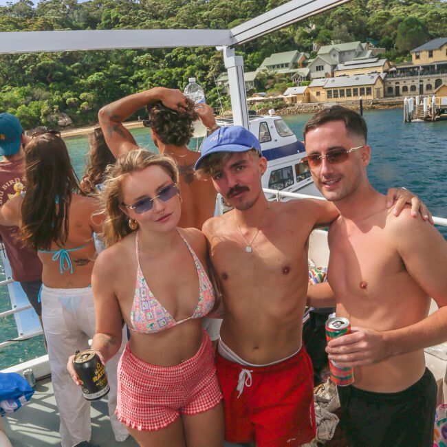 A group of young adults in swimwear pose on a boat with drinks in hand. They are smiling and enjoying a sunny day, with lush green trees and buildings visible onshore in the background.